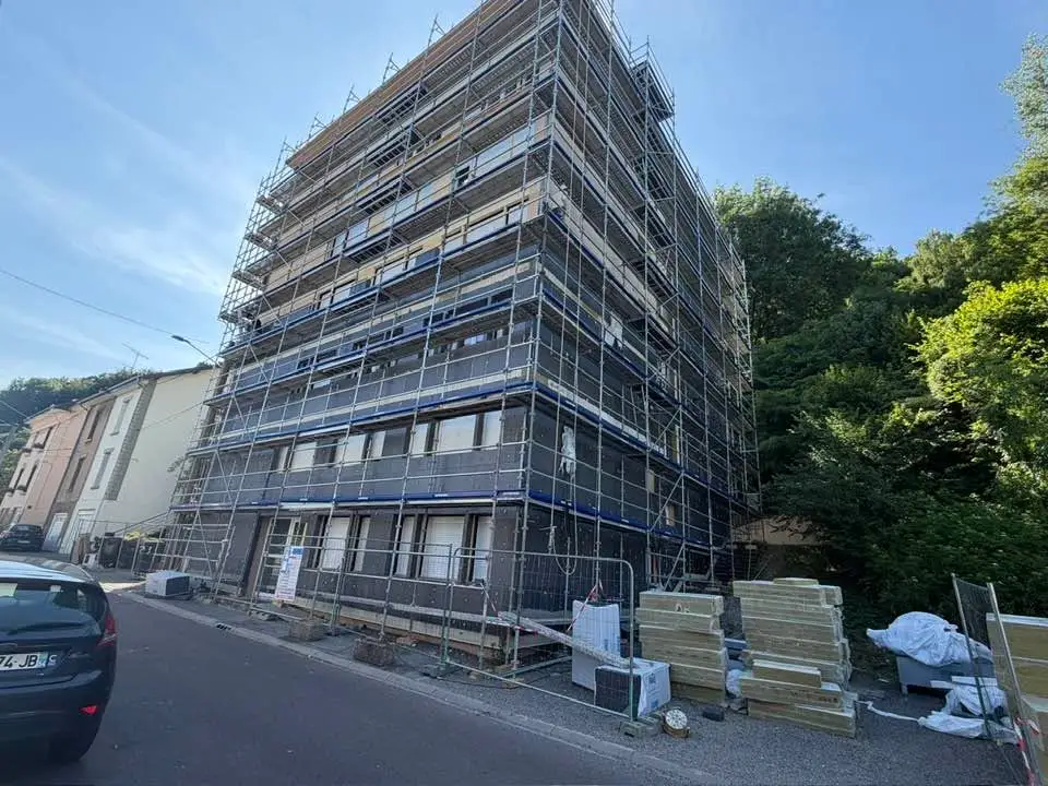 Façade d’immeuble entièrement échafaudée, paquets de panneaux isolants stockés au pied du bâtiment derrière des barrières.