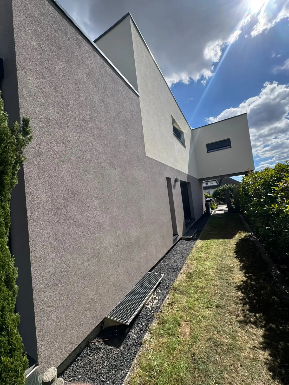 Vue rasante le long d’un mur latéral en enduit gris clair, bande de gravier et caniveau à grille en pied de mur.