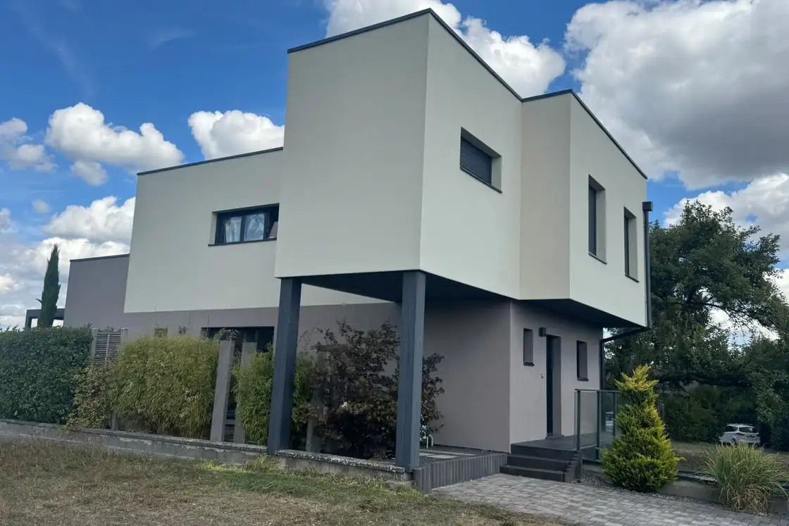 Façade latérale d’une maison contemporaine couverte d’un enduit de finition clair, menuiseries de teinte anthracite.