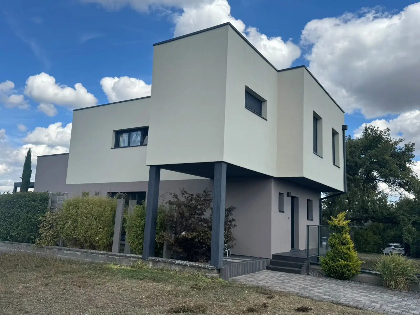 Angle de la maison, volume blanc cassé en porte-à-faux posé sur des poteaux sombres, niveau bas en enduit gris clair.