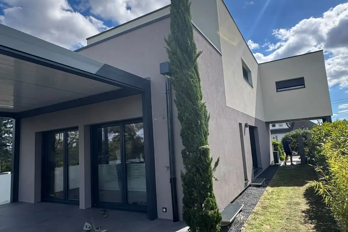 Terrasse couverte d’une maison contemporaine aux grandes baies vitrées, murs recouverts d’un enduit de finition clair.