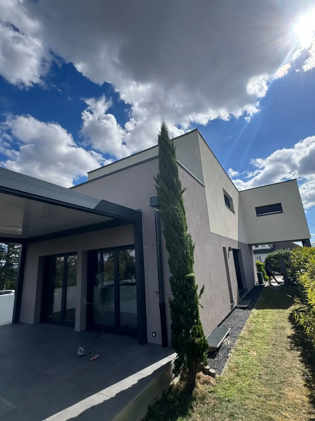Terrasse couverte par une pergola à lames, baies vitrées anthracite et murs en enduit gris clair.