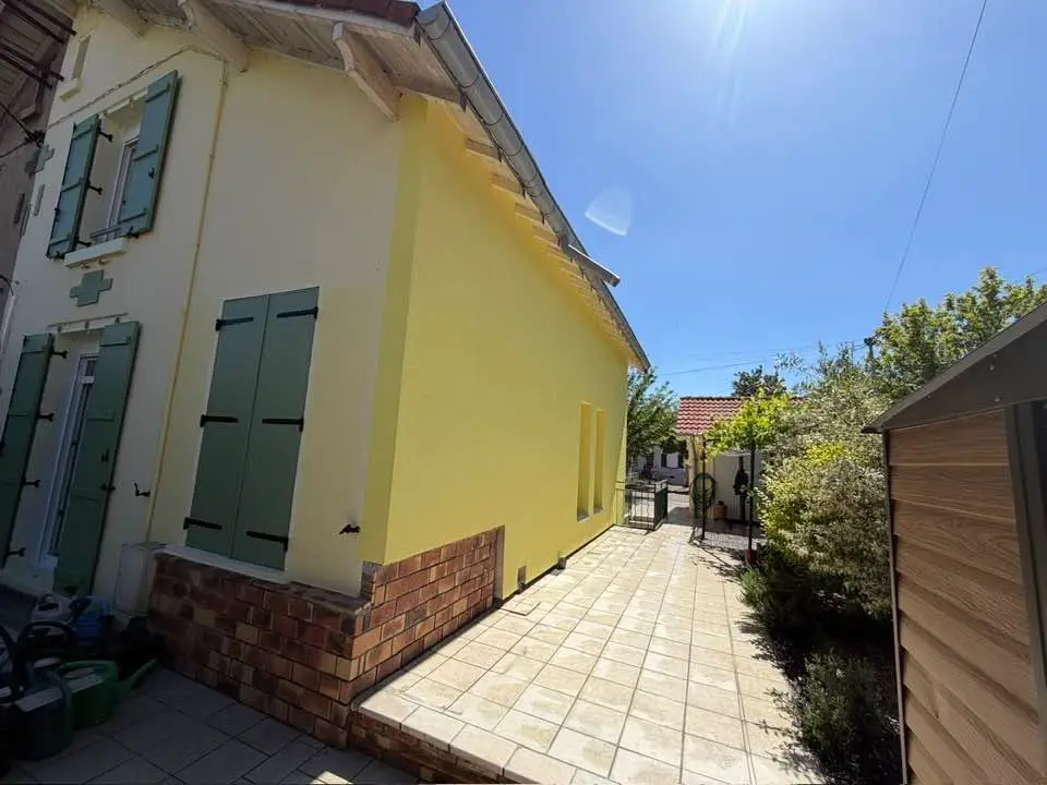 Pignon d’une maison après isolation par l’extérieur, enduit de finition jaune, soubassement en brique et volets verts.